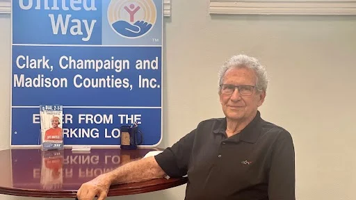 John Detrick is sitting in a chair, looking at the camera, United Way poster is in the background.